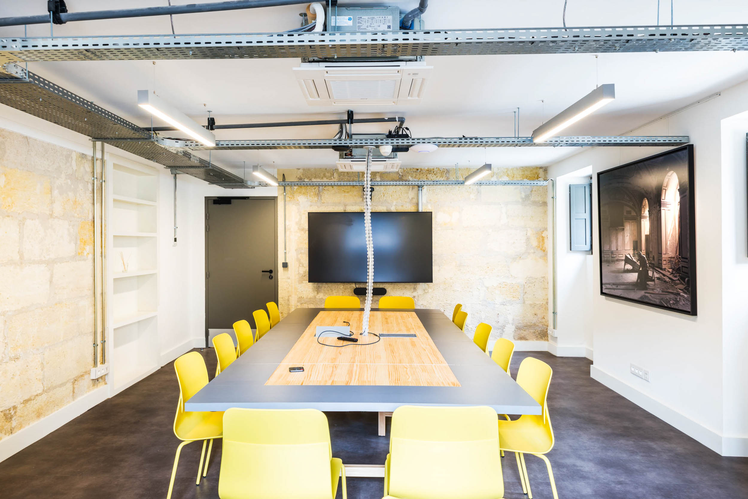 Une salle de conférence moderne, parfaite pour un photographe d'architecture et de décoration d'intérieur, dispose d'une longue table entourée de chaises jaune vif. La table est dotée d'une multiprise intégrée au centre. Un téléviseur à écran plat est fixé au mur en face de la porte, avec un éclairage de style industriel et des tuyaux apparents au-dessus. Des murs de couleur crème et des œuvres d'art encadrées complètent cet espace d'inspiration bordeaux.