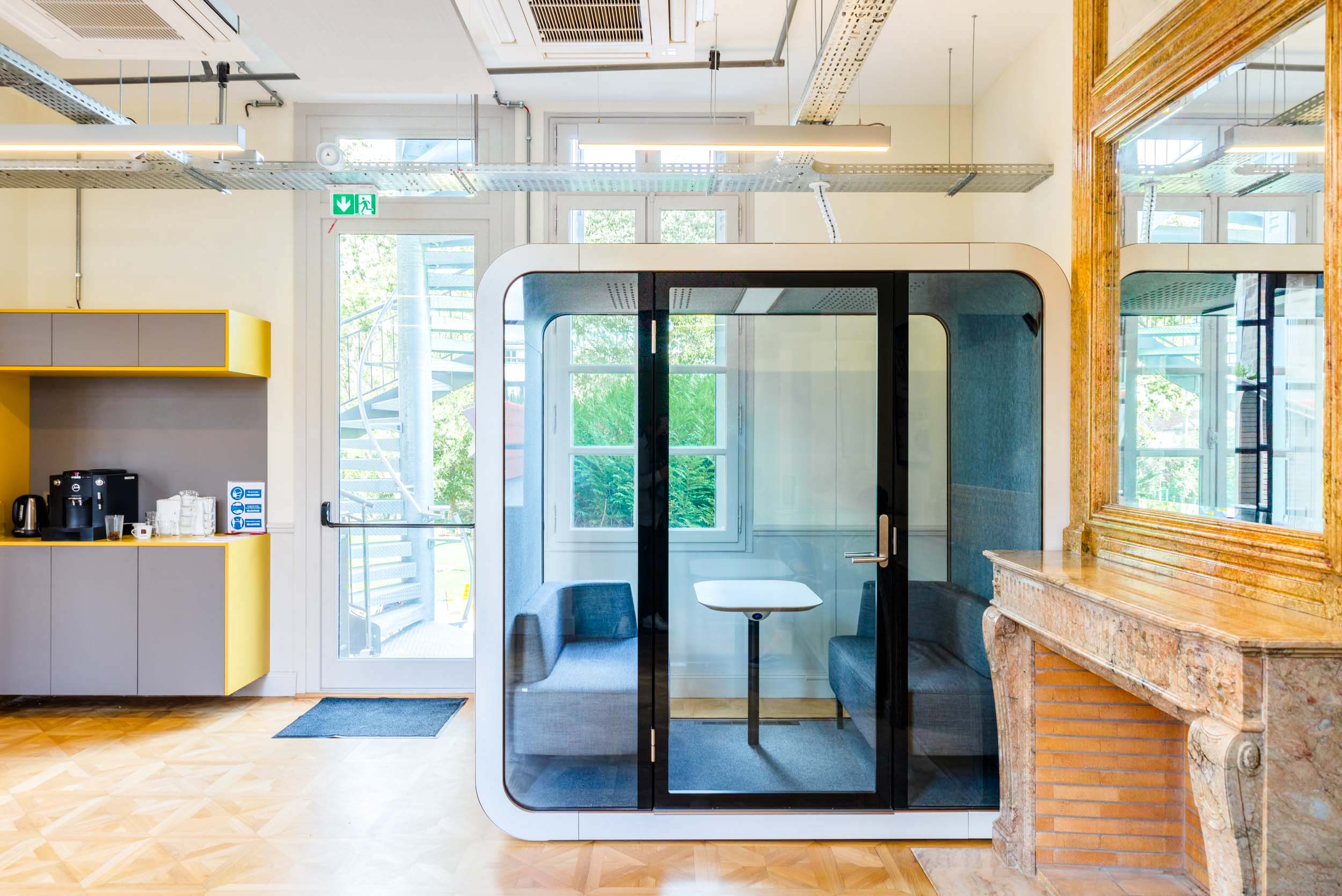 Un espace de bureau lumineux comprenant une cabine de réunion moderne aux parois vitrées avec des sièges et une petite table à l'intérieur. La cabine est située près d'une grande fenêtre et d'une cheminée avec un miroir décoratif au-dessus. À côté de la cabine se trouve un coin kitchenette gris et jaune avec une machine à café, parfait pour tout projet de décoration d'intérieur à Bordeaux.