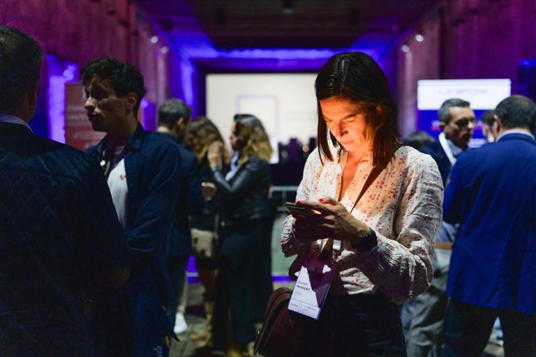 Une femme en chemisier blanc à fleurs, portant un cordon, se tient debout dans la salle de réception faiblement éclairée pendant la French Tech Night 2024, regardant son téléphone. Des personnes en tenue professionnelle sont rassemblées autour, engagées dans une conversation. L'arrière-plan a une teinte violette et un écran de présentation est visible au fond de la salle.