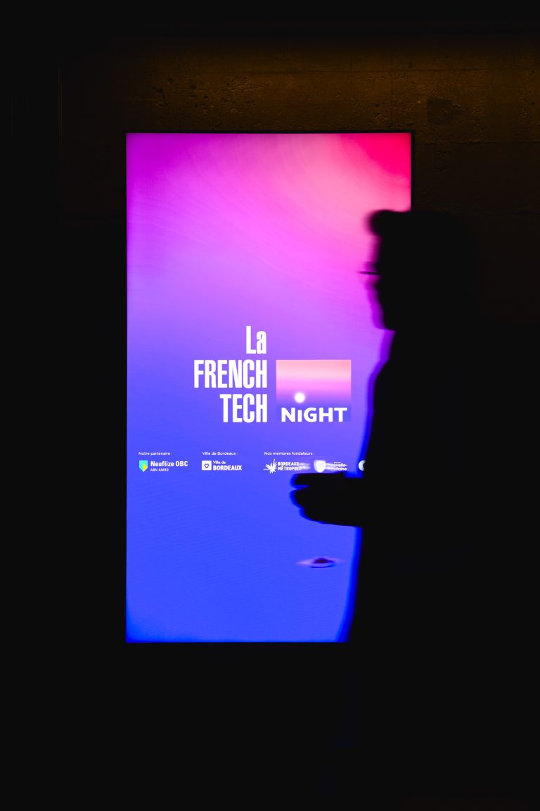 Une personne passe devant un panneau numérique éclairé sur lequel est écrit « French Tech Night 2024 » avec les logos des sponsors en dessous. Le panneau présente un fond dégradé passant du rose au violet. La scène est faiblement éclairée, ce qui fait apparaître la personne comme une silhouette sombre.