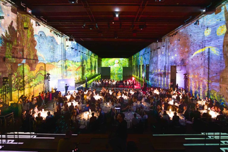 Un grand événement indoor, la French Tech Night 2024, réunit des personnes assises autour de tables rondes recouvertes de nappes blanches. Les murs et le plafond sont illuminés par des projections d'œuvres d'art animées et vibrantes, notamment des créations inspirées de Van Gogh. L'ambiance est animée, avec un éclairage chaleureux illuminant les tables.