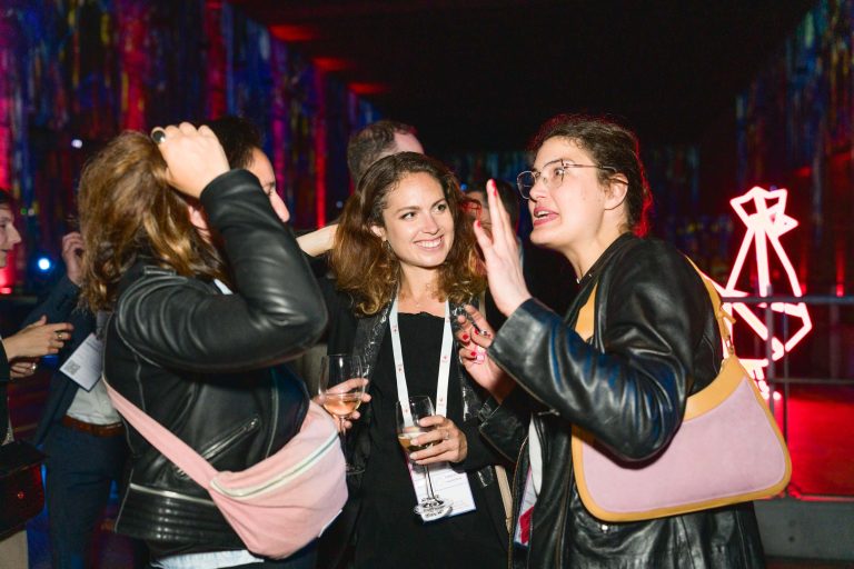 Un groupe de trois femmes participent à une conversation animée lors de ce qui semble être la French Tech Night 2024, avec un éclairage vibrant et coloré en arrière-plan. Elles portent des vêtements décontractés avec des vestes en cuir et portent des sacs à bandoulière. L'une tient un verre et une autre fait des gestes expressifs avec ses mains.