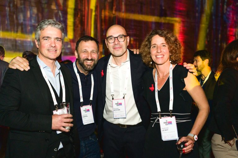 Quatre personnes se tiennent côte à côte à la French Tech Night 2024, souriant à la caméra. Chacun porte un badge nominatif et tient une boisson. L'arrière-plan présente un éclairage coloré et flou. Le groupe comprend trois hommes en tenue professionnelle et une femme en tenue noire.