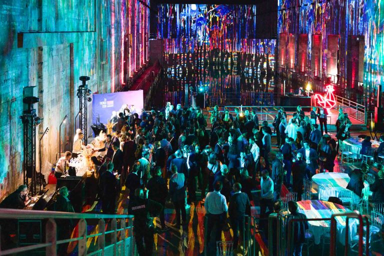 Une foule nombreuse se rassemble dans un lieu animé et illuminé pour la French Tech Night 2024, avec des lumières multicolores et des visuels projetés sur de hauts murs. Les gens participent à des conversations et à des activités à proximité de différentes stations. L'espace dégage une atmosphère énergique avec un décor artistique et moderne.