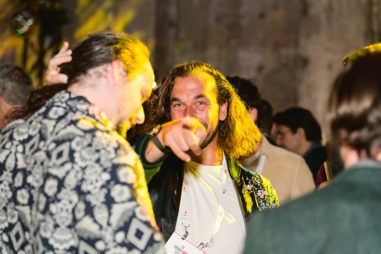 Deux individus interagissent lors de la French Tech Night 2024, un événement social animé. La personne au centre, vêtue d'une veste à motifs et d'une chemise blanche, sourit et pointe vers la caméra. La personne à gauche, vêtue d'une chemise à motifs noire et blanche, est légèrement détournée. L'arrière-plan est faiblement éclairé et d'autres participants sont visibles.