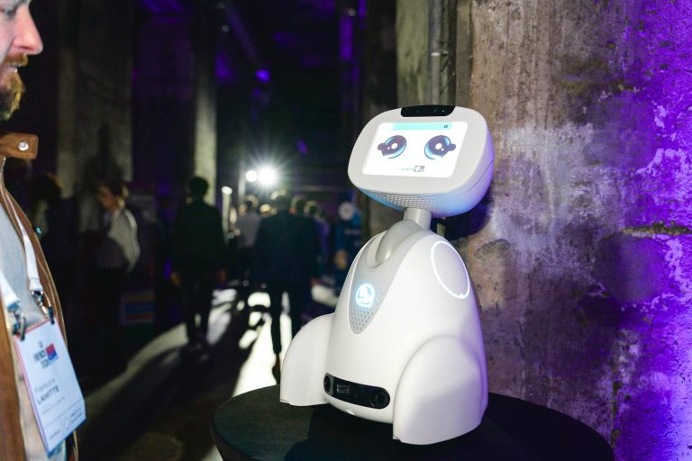 Lors de la French Tech Night 2024, un robot humanoïde blanc doté d'un écran affichant deux yeux se tient sur une table, baignée de lumières bleues et violettes. Des personnes se rassemblent dans l'arrière-plan faiblement éclairé de l'événement tandis qu'une personne avec un badge est visible sur le côté gauche, partiellement dans le cadre, regardant le robot.