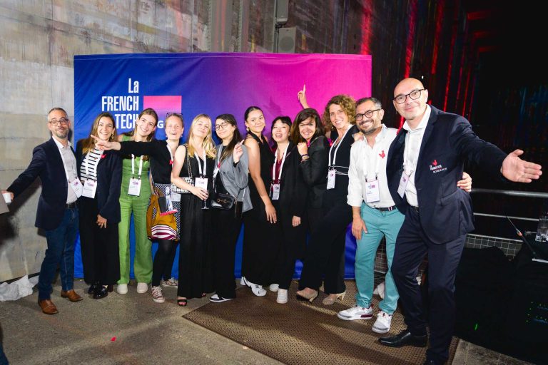 Un groupe de douze personnes se tient devant une bannière de La French Tech lors de la French Tech Night 2024. Ils sourient, certains font des gestes ludiques comme des signes de paix et pointent du doigt. La plupart portent des cordons de conférence et sont habillés de manière décontractée ou semi-formelle devant un mur industriel partiellement éclairé.