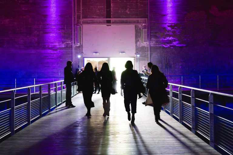 Des gens marchent sur un pont aux tons violets, la nuit. Leurs silhouettes projettent des ombres sur le sol éclairé. Plusieurs individus s'attardent sur les bords, et une porte avec une lumière blanche et chaude se trouve au bout du pont, suggérant l'entrée d'un bâtiment. Bienvenue à la French Tech Night 2024.