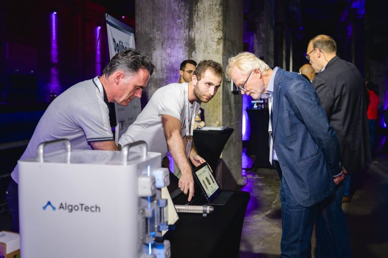 Trois hommes à la French Tech Night 2024 interagissent sur un stand intitulé « AlgoTech ». Deux hommes, l'un aux cheveux gris et l'autre aux cheveux noirs, se tiennent derrière le stand et font la démonstration d'une tablette à un troisième homme aux cheveux gris portant des lunettes et un blazer bleu. D'autres sont en arrière-plan, sous un éclairage violet tamisé.