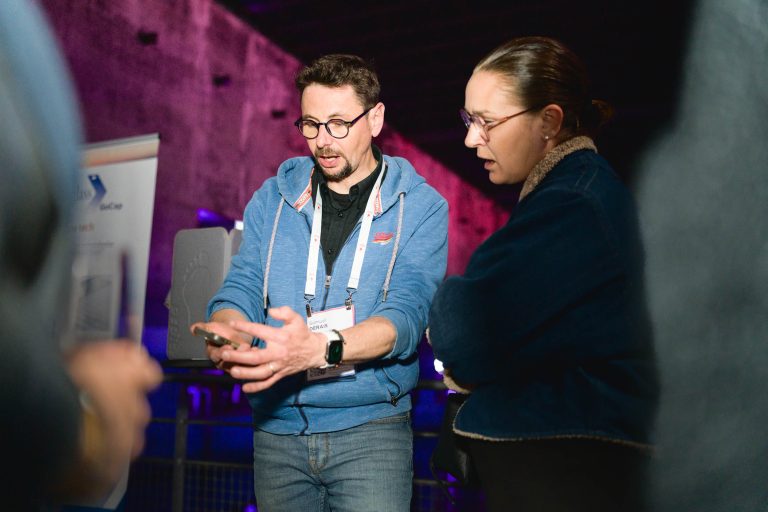 Deux individus discutent dans un intérieur faiblement éclairé lors de la French Tech Night 2024. La personne de gauche fait des gestes tout en tenant un petit appareil, peut-être un téléphone, et la personne de droite écoute attentivement, les mains le long du corps. Tous deux portent une tenue décontractée et des lunettes. Une bannière est partiellement visible en arrière-plan.