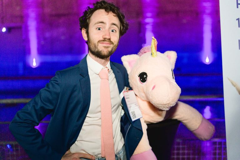 Un homme aux cheveux bouclés et à la barbe, vêtu d'une veste de costume bleue, d'une chemise blanche et d'une cravate rose, pose avec une grande licorne en peluche. Il porte un badge de conférence sur son côté droit et se trouve devant un arrière-plan flou éclairé en violet à la French Tech Night 2024.