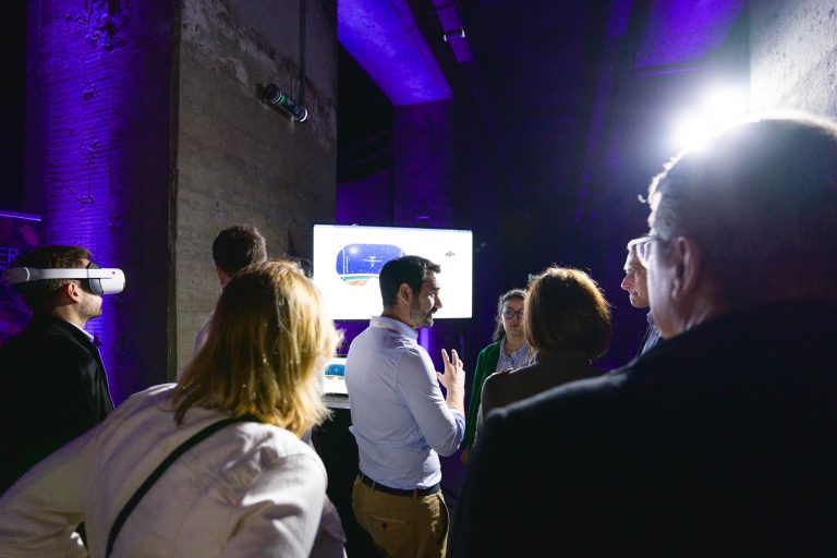 Un petit groupe de personnes se rassemble dans une salle faiblement éclairée par un éclairage ambiant violet pendant la French Tech Night 2024. Un homme au centre parle, fait des gestes avec ses mains, tandis qu'un écran affichant une présentation est visible en arrière-plan. Une personne porte un casque de réalité virtuelle, et d'autres écoutent attentivement.