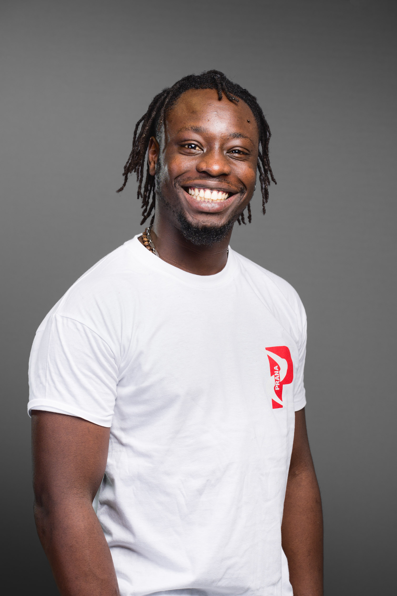 Une personne aux dreadlocks mi-longues sourit largement en regardant la caméra. Elle porte un t-shirt blanc avec un logo rouge sur le côté gauche de la poitrine. Le fond est un dégradé de gris uni, parfait pour les portraits professionnels à Bordeaux.