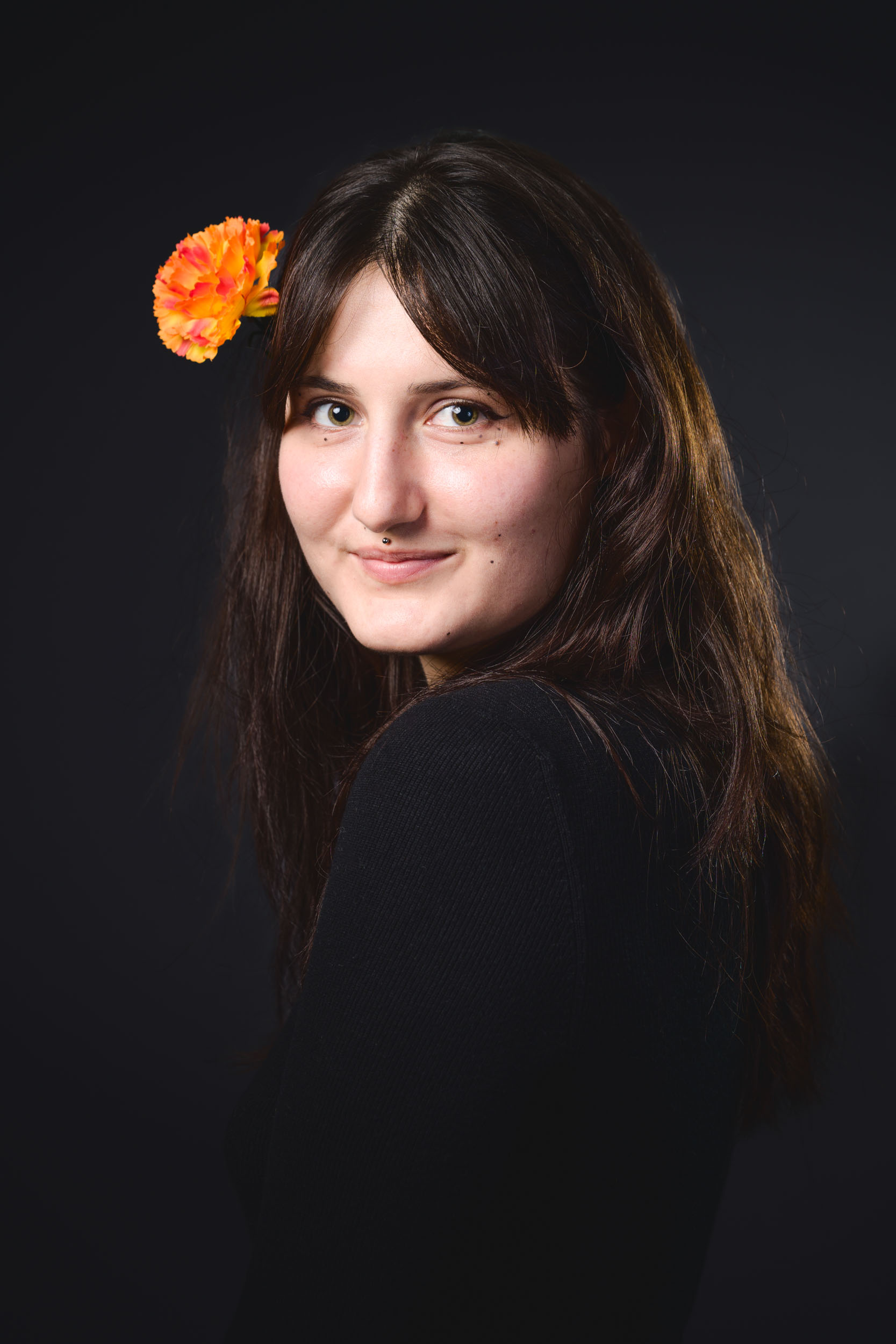 Une femme aux cheveux longs et bruns, au sourire léger, pose sur un fond sombre. Elle porte un haut noir et a une fleur d'œillet orange coincée dans ses cheveux au-dessus de son oreille gauche. Son regard est dirigé vers l'objectif, incarnant l'essence des portraits professionnels à Bordeaux.