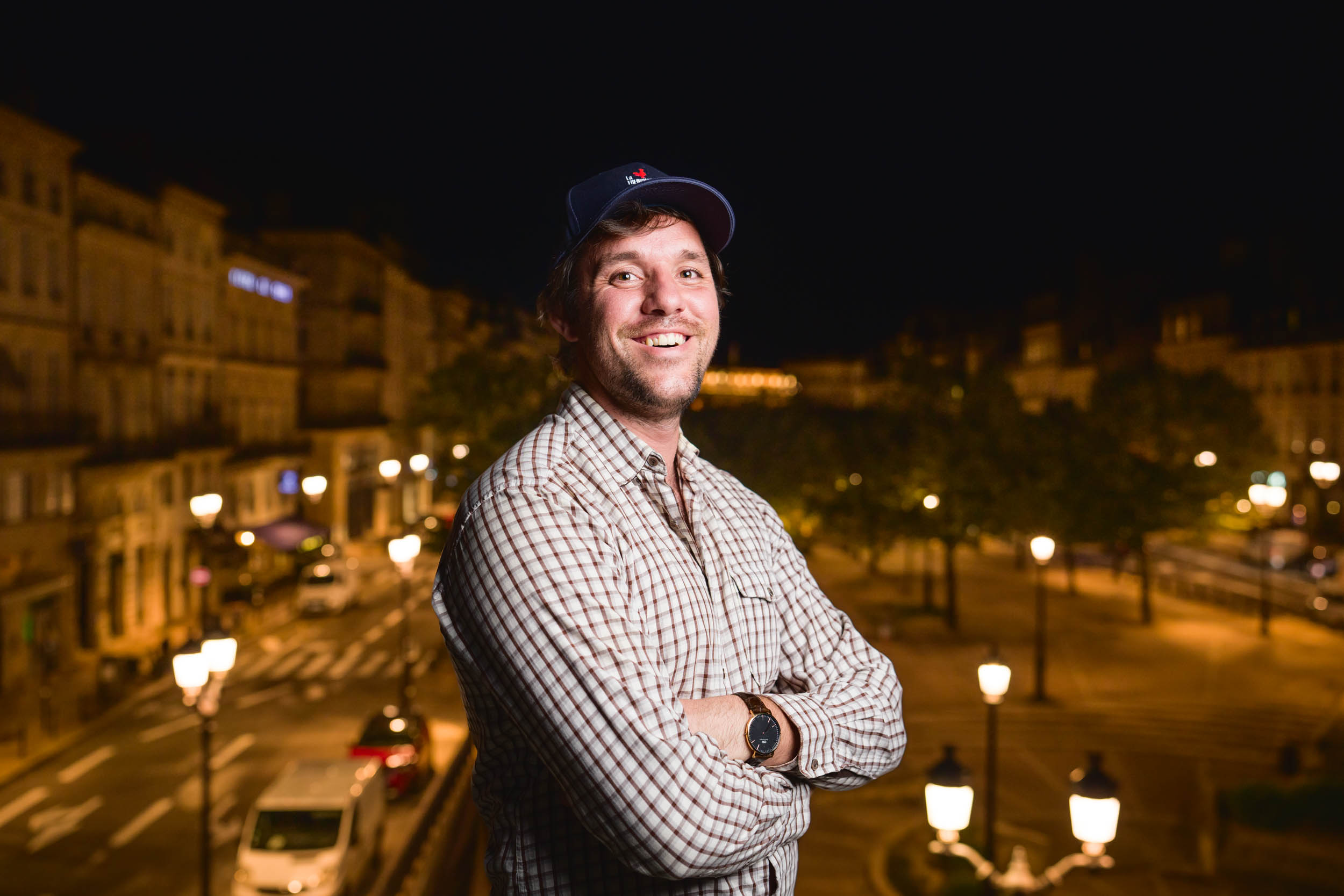 Un homme portant une casquette de baseball bleu marine et une chemise à carreaux sourit en se tenant debout, les bras croisés. Il est dehors la nuit avec des lampadaires et des bâtiments flous en arrière-plan. La scène est chaleureusement éclairée, donnant une atmosphère chaleureuse au décor nocturne, comme des portraits professionnels à Bordeaux.