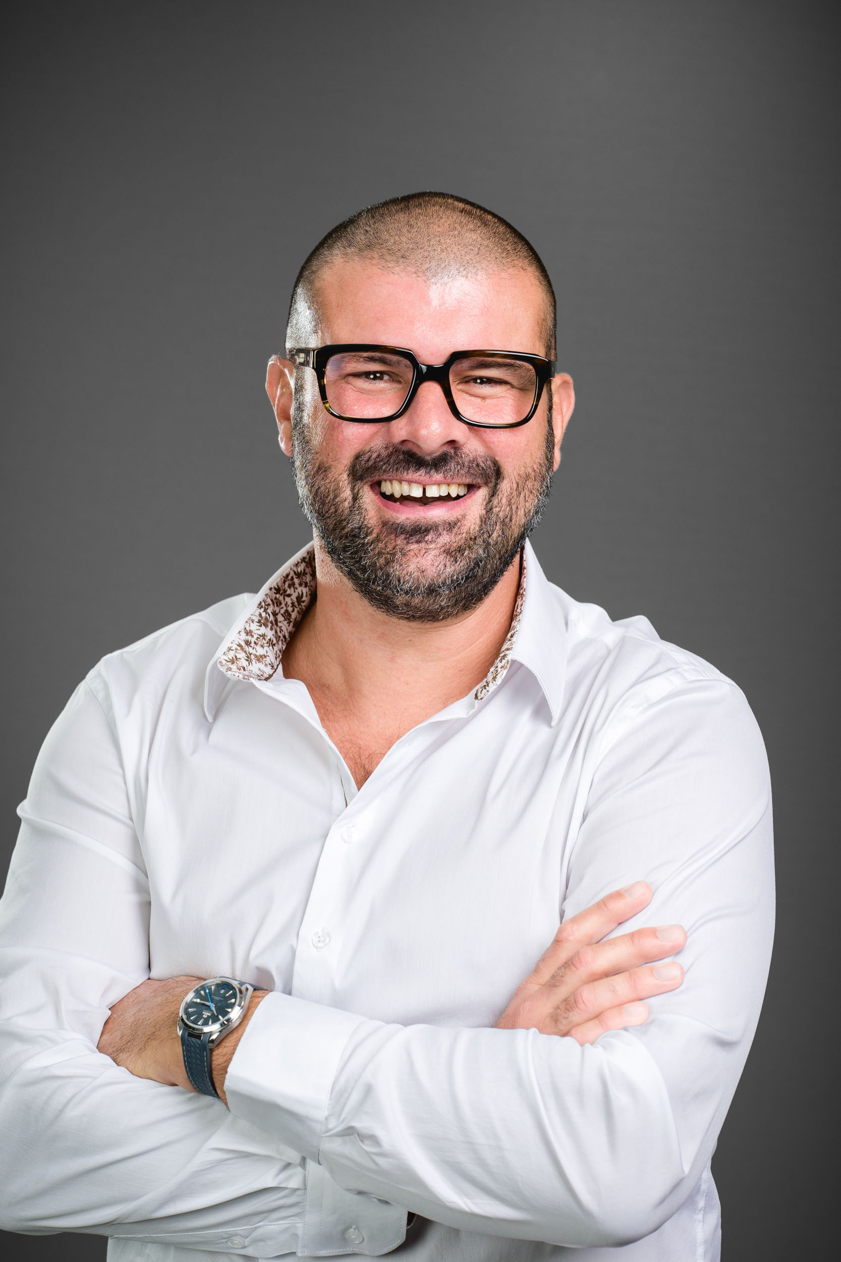 Un homme chauve avec une barbe et des lunettes à monture noire sourit largement. Il porte une chemise blanche aux manches retroussées, révélant un col intérieur à motifs. Ses bras sont croisés, laissant apparaître sa montre-bracelet au poignet gauche. Le fond de ce portrait professionnel à Bordeaux est un gris foncé uni.