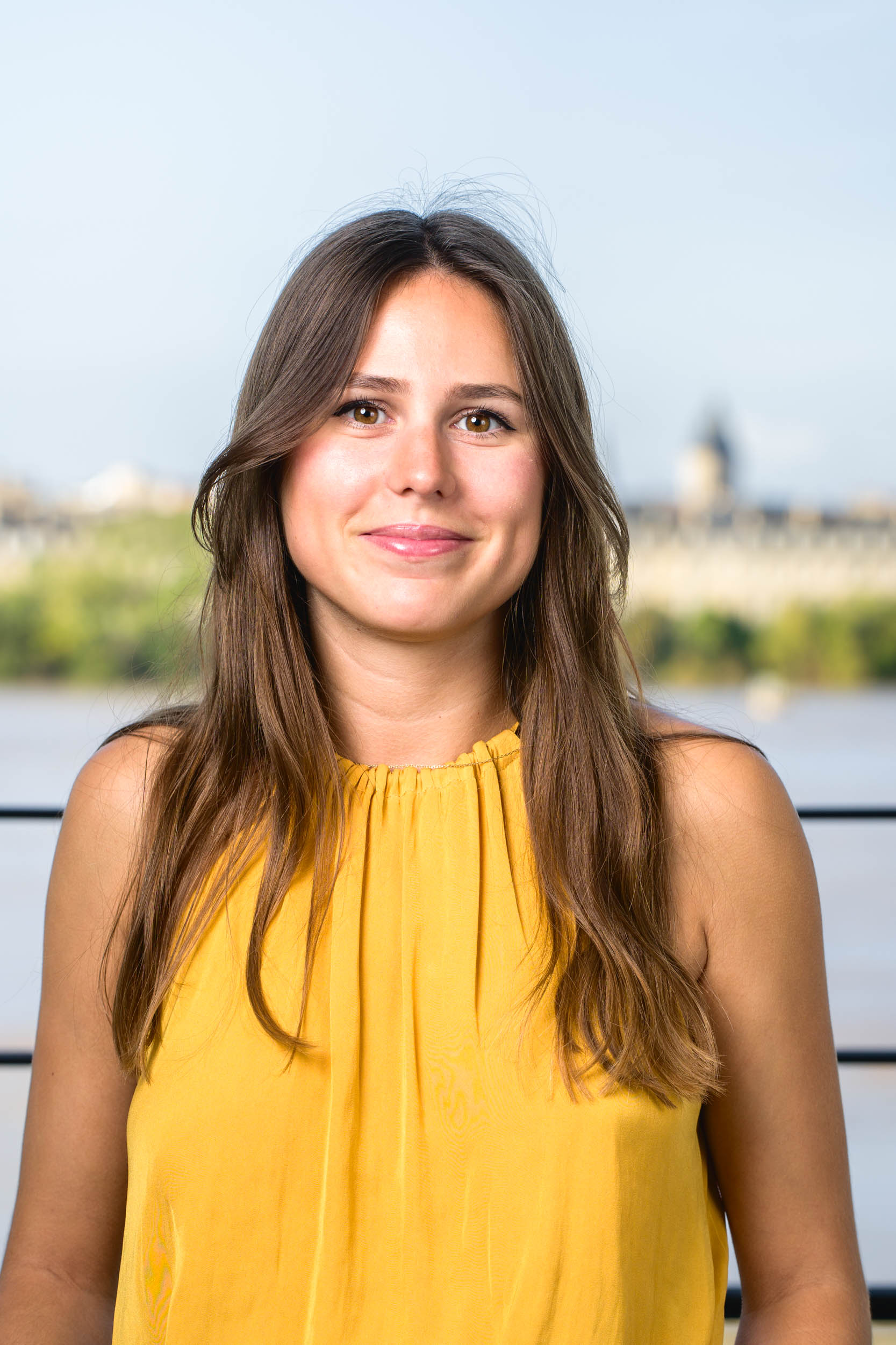 Une jeune femme aux longs cheveux bruns, souriante et tournée vers l'objectif, porte un haut jaune sans manches. L'arrière-plan présente un décor extérieur flou avec un plan d'eau et de la verdure, rappelant les portraits professionnels à Bordeaux. Le ciel est clair, annonçant une journée ensoleillée.