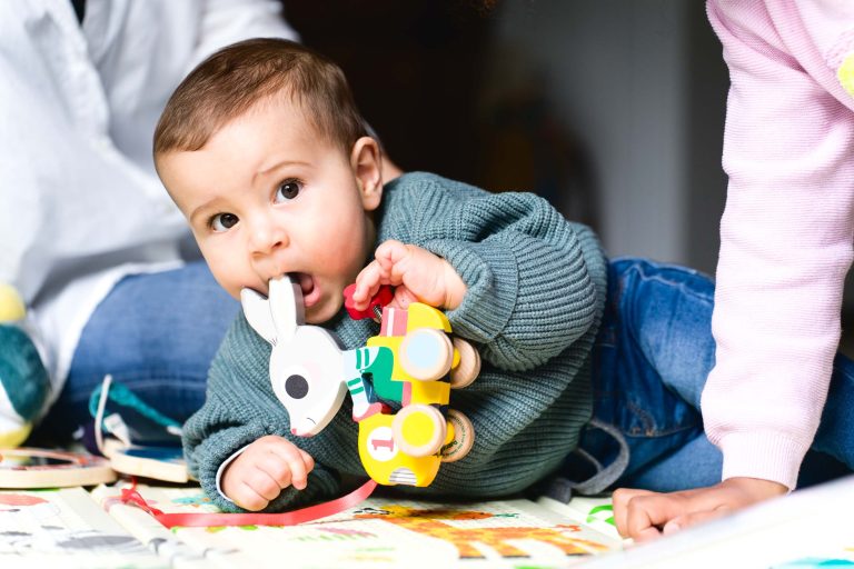 Un bébé est allongé sur le ventre sur un tapis de jeu coloré, vêtu d'un pull vert et d'un jean bleu. Le bébé tient un jouet en bois en forme de cheval à roulettes et en met une partie dans sa bouche. A proximité, un adulte et un autre enfant sont partiellement visibles lors de cette charmante séance photo famille Bordelaise.