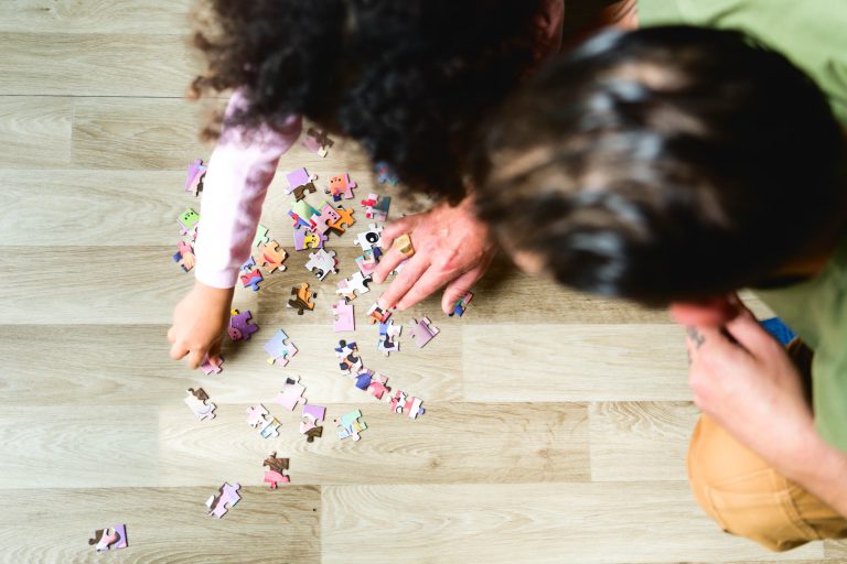 Deux individus sont vus d'en haut, en train d'assembler un puzzle coloré sur un plancher en bois clair. L'une des personnes a les cheveux bouclés et l'autre les cheveux raides et foncés. La scène capture un moment d'activité et de concentration partagée, rappelant une séance photo de la famille Bordelaise alors qu'ils assemblent les pièces du puzzle.