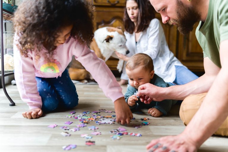 Une famille est réunie par terre pour assembler un puzzle. Une jeune fille en pull rose et jean bleu attrape les pièces tandis qu'un bébé regarde avec curiosité. Un homme adulte aide le bébé. En arrière-plan, une femme adulte et un chien observent, capturant un instantané de cette séance photo de la famille Bordelaise.