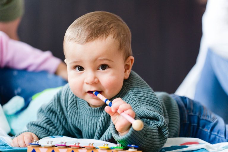 Un bébé aux cheveux châtains courts, vêtu d'un pull vert, est allongé sur un tapis de jeu coloré. Le bébé tient dans sa bouche un maillet de xylophone et regarde directement l'objectif. Des figures et des objets flous et hors de vue apparaissent à l'arrière-plan de cette séance photo de la famille Bordelaise.