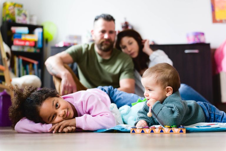 Une jeune fille est allongée sur le sol et sourit à l'objectif, tandis qu'un bébé à proximité tient un jouet et la regarde. En arrière-plan, un homme et une femme sont assis l'un à côté de l'autre, légèrement flous, avec des objets colorés et des jouets éparpillés un peu partout. Cette photo réconfortante de la séance de la famille Bordelaise capture magnifiquement leur joie détendue.