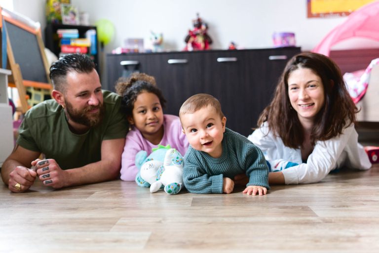 Sur cette photo de séance de la famille Bordelaise, une famille de quatre personnes est allongée sur le plancher en bois d'une salle de jeux. Le père barbu est à gauche, la jeune fille aux cheveux bouclés est à ses côtés, le bébé en pull gris est au centre et la mère aux cheveux longs est à droite. Des jouets et des armoires sont visibles à l'arrière-plan.