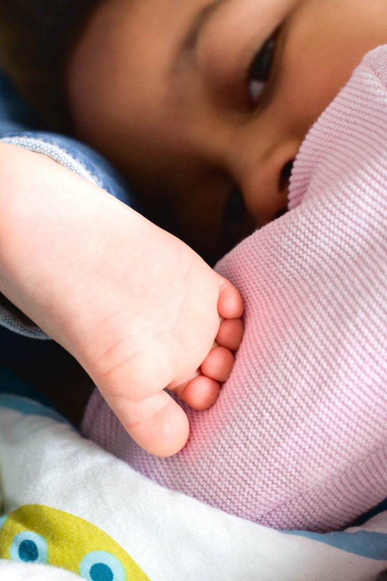 Une image en gros plan d'un bébé allongé, partiellement recouvert d'une couverture texturée rose, évoque la chaleur d'une séance photo de la famille Bordelaise. Le pied du bébé est au premier plan, placé près de son visage. La peau lisse et l'éclairage doux créent une atmosphère chaleureuse tandis que la tête du bébé dépasse de la couverture.