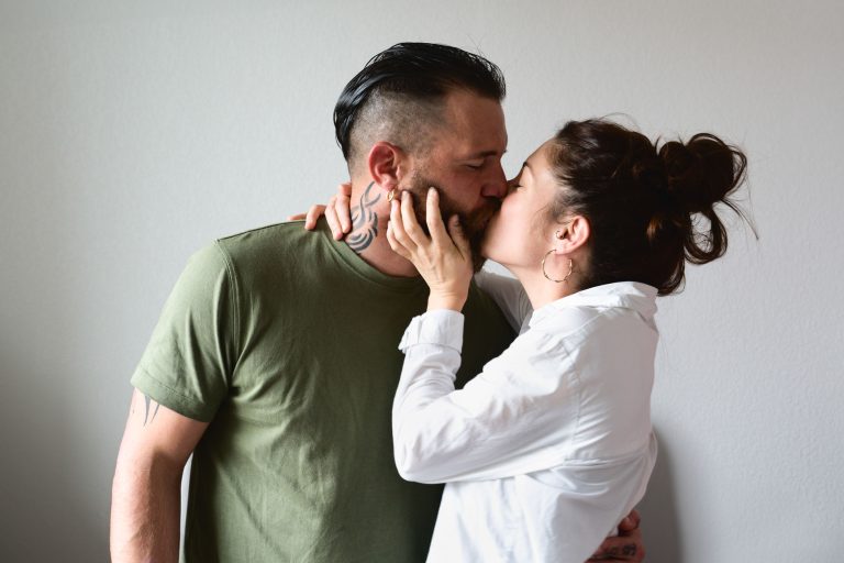 Un homme barbu, vêtu d'un t-shirt vert, et une femme aux cheveux en chignon, vêtue d'une chemise blanche, s'embrassent. L'homme passe son bras autour de la femme tandis qu'elle lui touche doucement le visage avec sa main gauche. Ce moment intime est capturé lors de leur séance photo famille Bordelaise devant un mur uni de couleur claire.