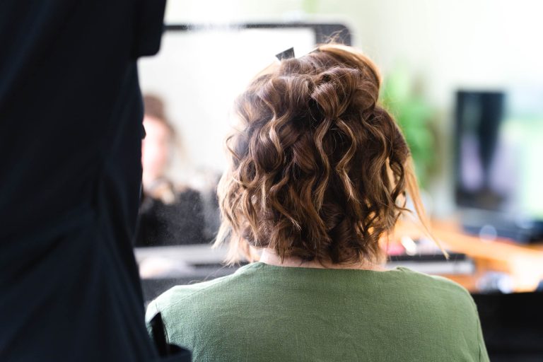 Aline aux cheveux bruns courts et bouclés, vêtue d’une chemise verte, est assise de dos devant un miroir pendant qu’une coiffeuse vaporise un produit sur sa coiffure, le reflet dans le miroir restant volontairement flou pour mettre l’accent sur le geste de préparation.