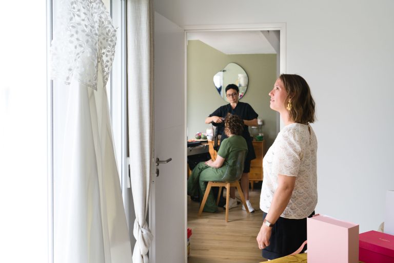 Une témoin en haut en dentelle blanche se tient près d’une grande fenêtre lumineuse et contemple sa robe de mariée en dentelle suspendue à une porte, tandis qu’au fond, dans une autre pièce visible par l’embrasure, Alinese fait maquiller et une autre apparaît dans le reflet d’un miroir rond.