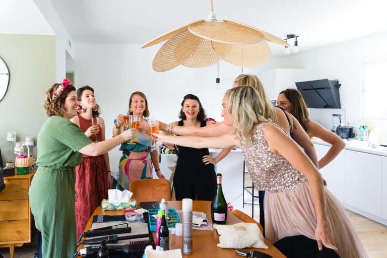 Sept proches d’Aline se tiennent autour d’une table dans une cuisine lumineuse à Montussan, souriant et trinquant avec des verres de vin mousseux, vêtues de robes de fête et de bandeaux fleuris, entourées de pâtisseries, de bouteilles et de produits de coiffure posés sur le plan de travail.