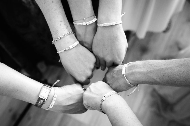 Cinq mains féminine ornées de bracelets en perles et d’une montre se rejoignent pour un check du poing au-dessus d’un plancher en bois, dans une photo en noir et blanc prise en plongée qui symbolise la complicité et l’unité entre les témoins d'Aline. ​