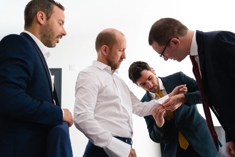 Le marié Yohan, au centre en gilet bleu marine et nœud papillon fleuri, ajuste son nœud papillon les yeux fermés tandis que trois témoins en costume l'entourent pour vérifier sa tenue devant un fond blanc épuré, créant un moment complice et concentré de préparation juste avant la cérémonie de son mariage avec Aline.