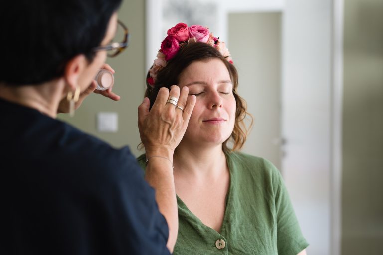 Chrystelle Bize, maquilleuse professionnelle aux cheveux bruns ondulés et lunettes, applique au pinceau un fard à paupières rosé sur les yeux fermés d’Aline, assise en robe verte dans une pièce lumineuse aux murs clairs et porte en arrière‑plan.