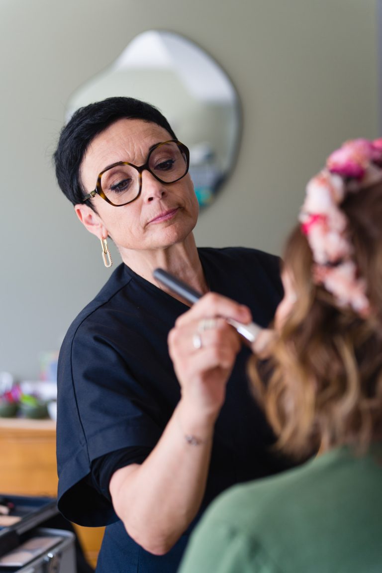 La maquilleuse professionnelle Chrystelle Bize, aux cheveux bruns courts, portant des lunettes et un haut noir, applique au pinceau le maquillage sur le visage d’Aline coiffée d’un bandeau floral rose, toutes deux penchées l’une vers l’autre dans une pièce lumineuse, créant une atmosphère intime de préparation avant la cérémonie. ​