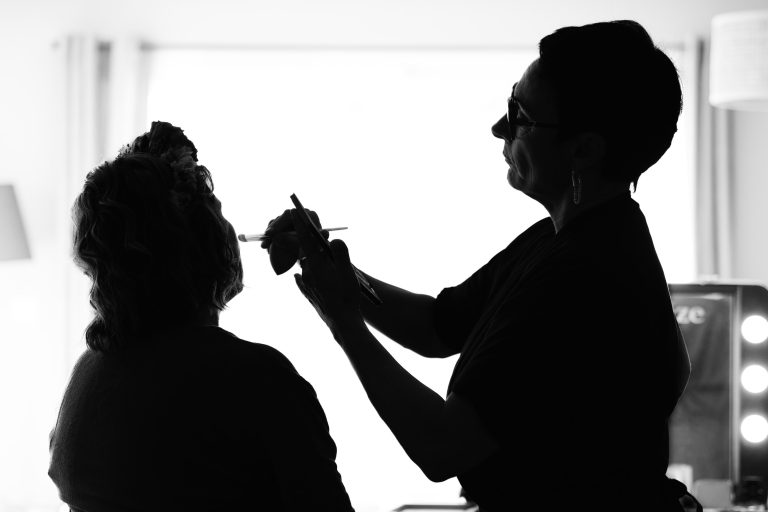 Une maquilleuse en silhouette se penche vers Aline assise pour lui maquiller le visage, tenant une palette et un pinceau, toutes deux de profil devant une grande fenêtre très lumineuse qui diffuse une forte lumière blanche et dessine leurs contours.