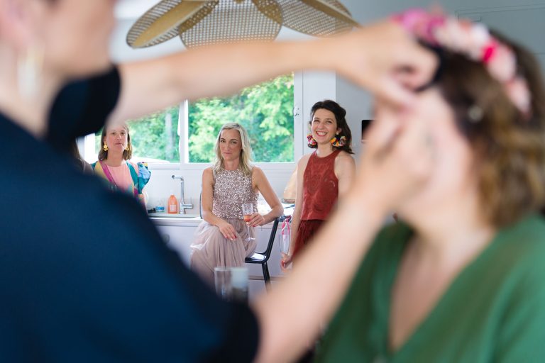 Trois témoins se tiennent dans une pièce lumineuse pendant les préparatifs du mariage d’Aline, deux souriantes et une au visage plus neutre, tandis qu’au premier plan flou une maquilleuse travaille sur une personne hors champ, sous un grand luminaire moderne avec vue sur la verdure extérieure. ​