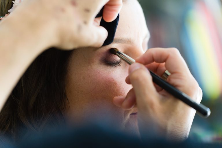 Gros plan sur le visage d’Aline aux yeux fermés pendant que la maquilleuse applique au pinceau un fard à paupières brun sur sa paupière, sous une lumière douce qui met en valeur les détails de son maquillage de mariée, l’arrière‑plan restant flou.