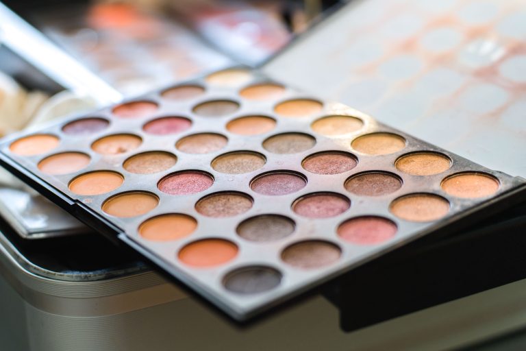 Gros plan sur une palette de maquillage ouverte posée sur une table, composée de fards à paupières ronds dans des tons bruns, dorés, pêche et rose, avec des pinceaux flous en arrière‑plan, utilisée pour préparer Aline avant son mariage.