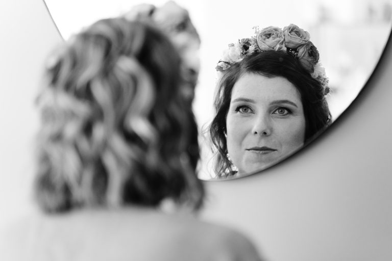 Aline aux cheveux ondulés porte une couronne de fleurs et une robe de mariée en dentelle, observant attentivement son reflet dans un miroir rond accroché à un mur clair, dans une scène en noir et blanc où son dos flou encadre nettement son visage. ​