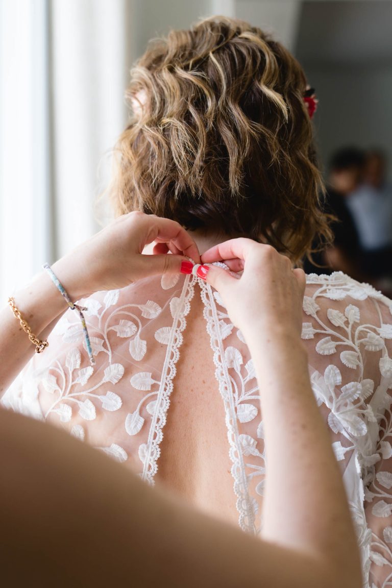 Gros plan sur le dos d’Aline en robe de mariée blanche en dentelle florale, tandis qu’une proche referme patiemment la rangée de petits boutons nacrés sous une lumière naturelle douce qui met en valeur les détails du tissu. ​