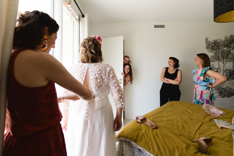 Aline en robe de mariée blanche se tient près d’une fenêtre pendant qu’une femme attache les boutons dans son dos, tandis que trois proches souriants l’observent depuis l’embrasure de la porte d’une chambre où l’on voit un lit à couvre‑lit jaune sur lequel sont posées ses chaussures et des accessoires roses.