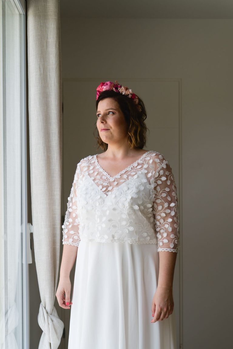 Aline en robe de mariée en dentelle blanche à manches trois quarts se tient près d’une grande fenêtre aux rideaux légers, coiffée d’un bandeau floral rose, regardant vers l’extérieur tandis qu’une douce lumière naturelle éclaire son profil.