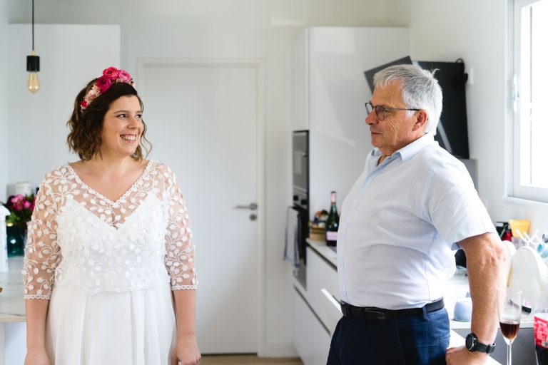 Aline en robe de mariée blanche aux manches en dentelle et couronne de fleurs roses sourit dans une cuisine lumineuse en discutant avec son père, un homme plus âgé portant des lunettes et une chemise bleu clair, tandis que la lumière du soleil traverse la fenêtre et crée une atmosphère chaleureuse. ​