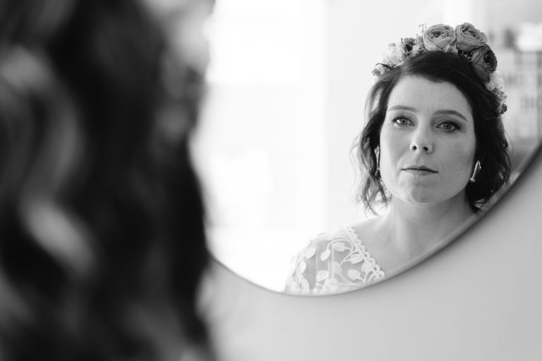 Aline aux cheveux ondulés, portant une couronne de fleurs et une robe de mariée en dentelle, se regarde dans un miroir rond accroché au mur, son visage calme et concentré éclairé en douceur dans une scène en noir et blanc, avec un avant‑plan légèrement flou suggérant la préparation du mariage. ​
