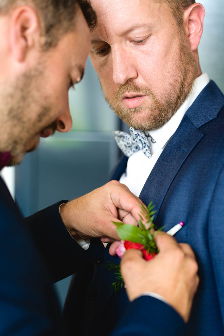 Yohan en costume bleu avec nœud papillon à motif floral se tient de profil pendant qu’un témoin en chemise blanche et bretelles lui épingle une boutonnière composée de verdure et d’une fleur rose sur le revers de sa veste, dans une pièce intérieure à l’arrière‑plan flou.