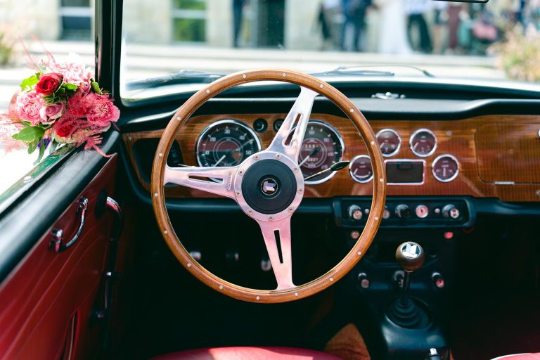 Gros plan sur le volant en bois et le tableau de bord d’une Triumph TR4A décapotable verte, avec ses cadrans et indicateurs analogiques sur fond de sellerie rouge, un bouquet de fleurs rouges et roses posé à gauche dans l’habitacle. ​