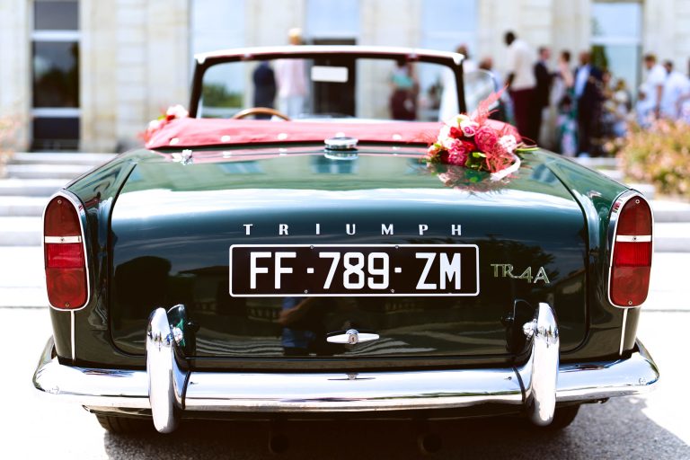 Triumph TR4A décapotable verte classique décorée d’un bouquet de fleurs rouges et roses sur le capot, garée devant la façade en pierre claire de la mairie de Montussan baignée de soleil, tandis que quelques invités flous se tiennent près de l’entrée.