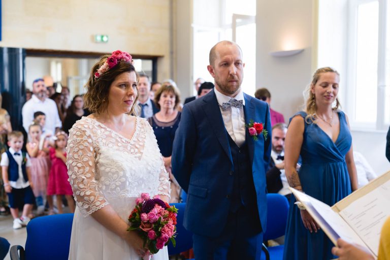 Aline en robe de mariée en dentelle blanche tenant un bouquet coloré se tient aux côtés de Yohan en costume bleu et nœud papillon dans la salle des mariages de la mairie de Montussan, tandis qu’une demoiselle d’honneur en robe bleue et plusieurs invités, dont des enfants, se rassemblent autour d’eux dans la lumière naturelle.